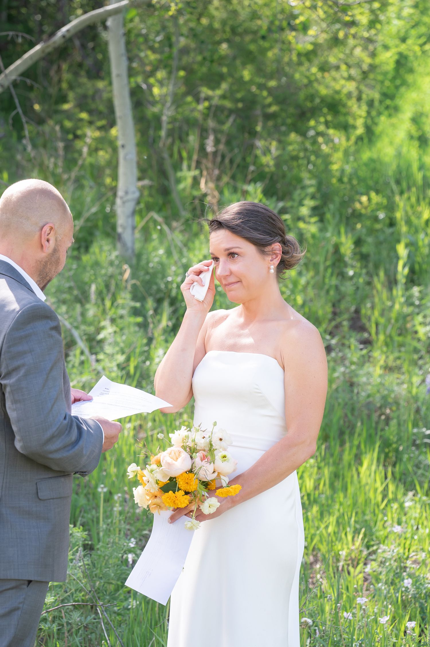 Emotional bride wiping tears holding yellow floral bouquet during outdoor wedding ceremony with groom reading vows.