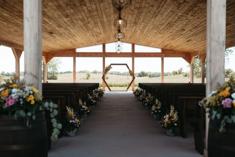 Rustic wedding chapel with wooden pews, floral arrangements, hexagonal arch, and scenic countryside views through open walls.