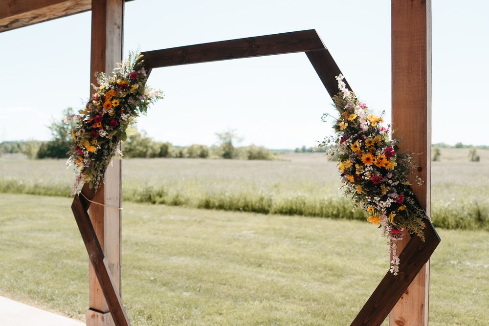 Hexagonal wooden wedding arch adorned with colorful wildflowers, set against a scenic green field backdrop.