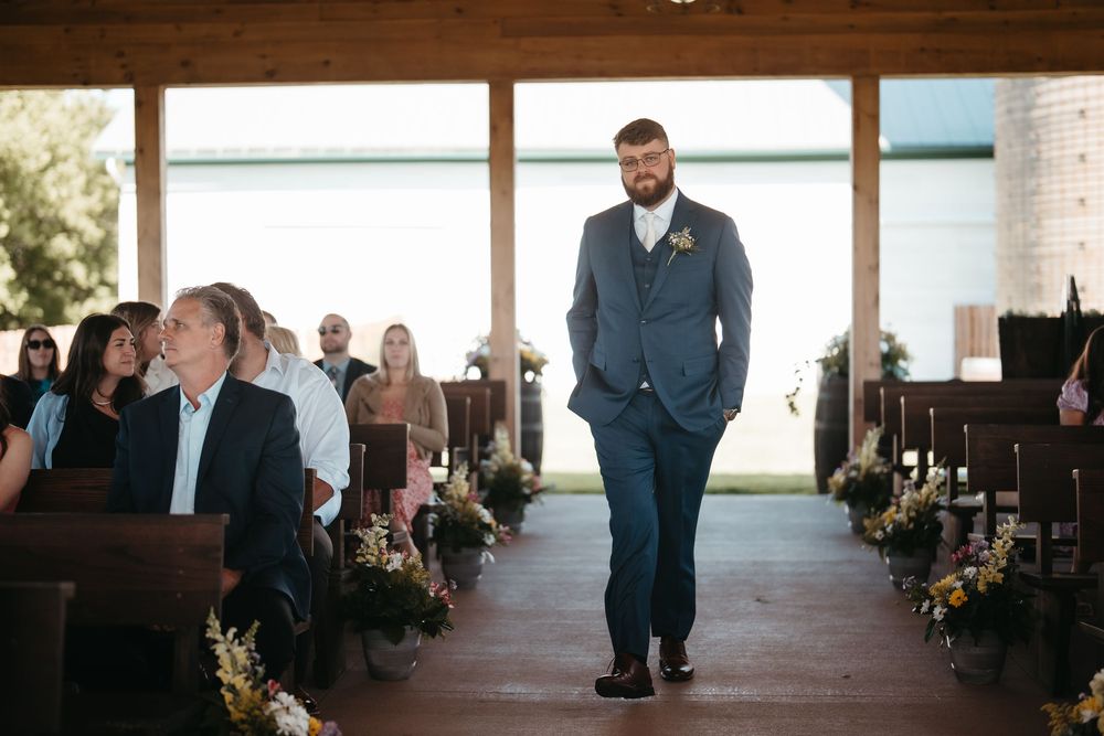 Groom in navy suit walks down wedding aisle in rustic chapel with wooden pews and floral decorations.