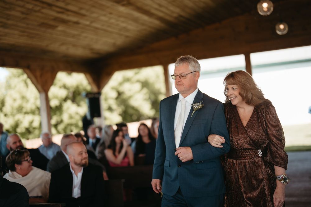 Couple walking arm in arm at an outdoor wedding ceremony under a wooden pavilion with guests seated nearby.