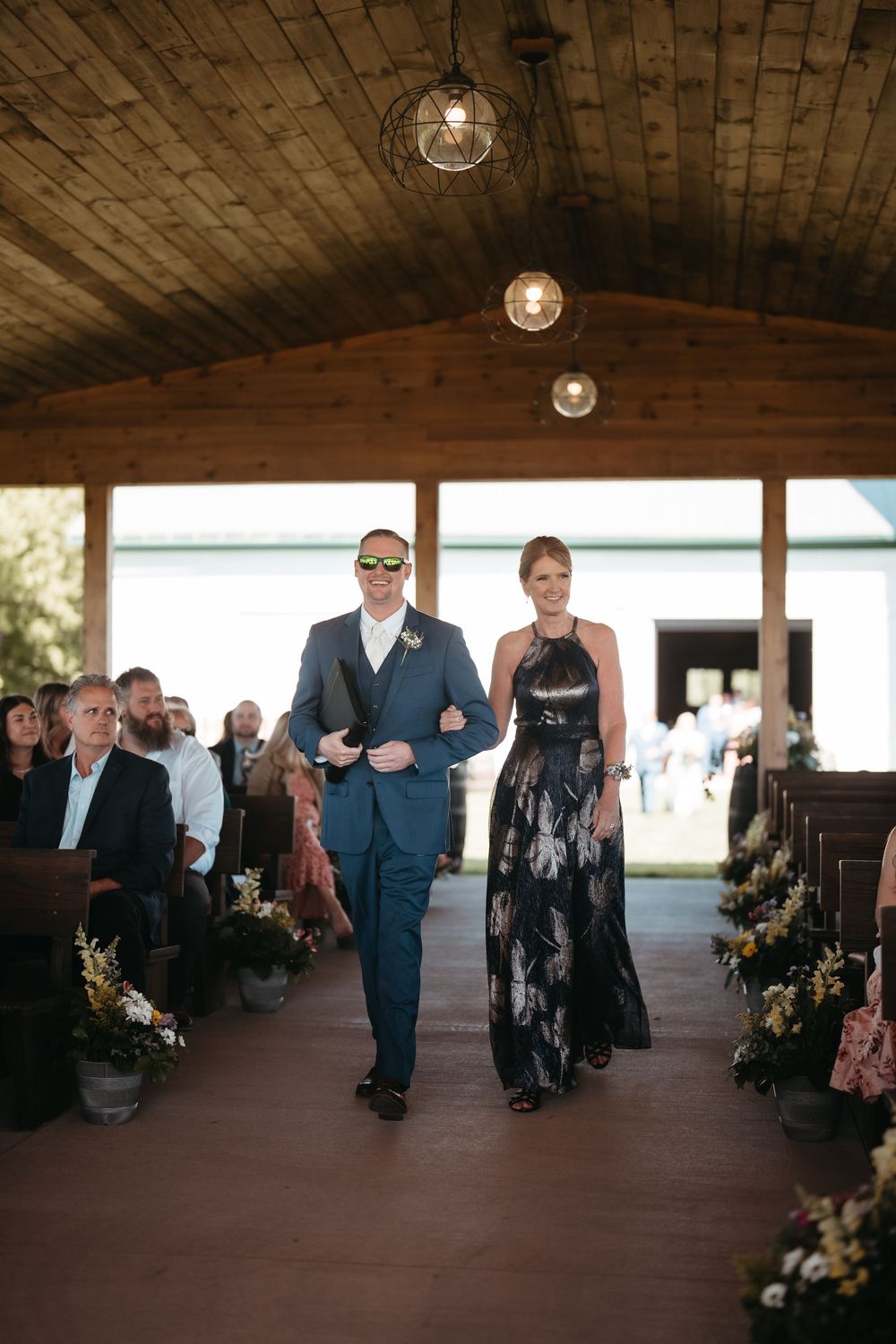 A groomsman in a blue suit escorts a bridesmaid in a floral gown down the aisle at a rustic barn wedding.