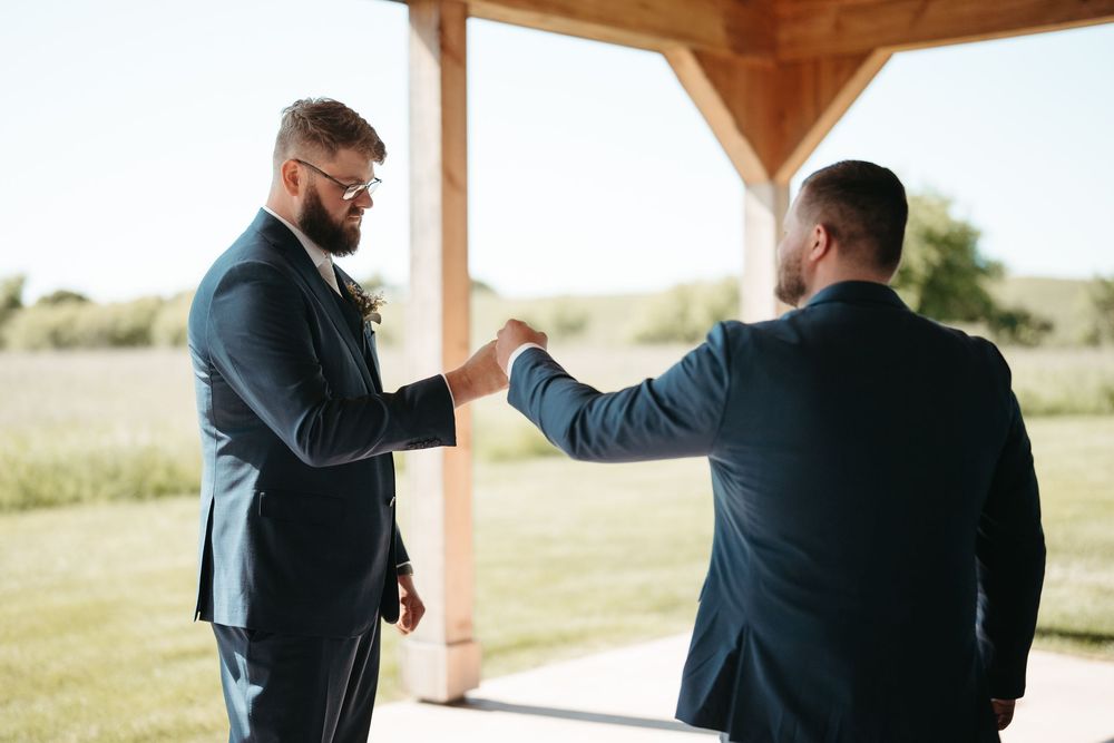 Two grooms in navy suits share a fist bump under a wooden pavilion at an outdoor wedding ceremony.