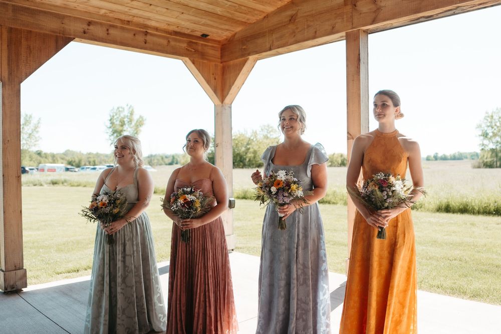 Four bridesmaids in mismatched floral and colorful maxi dresses holding bouquets under a wooden pavilion at an outdoor wedding.