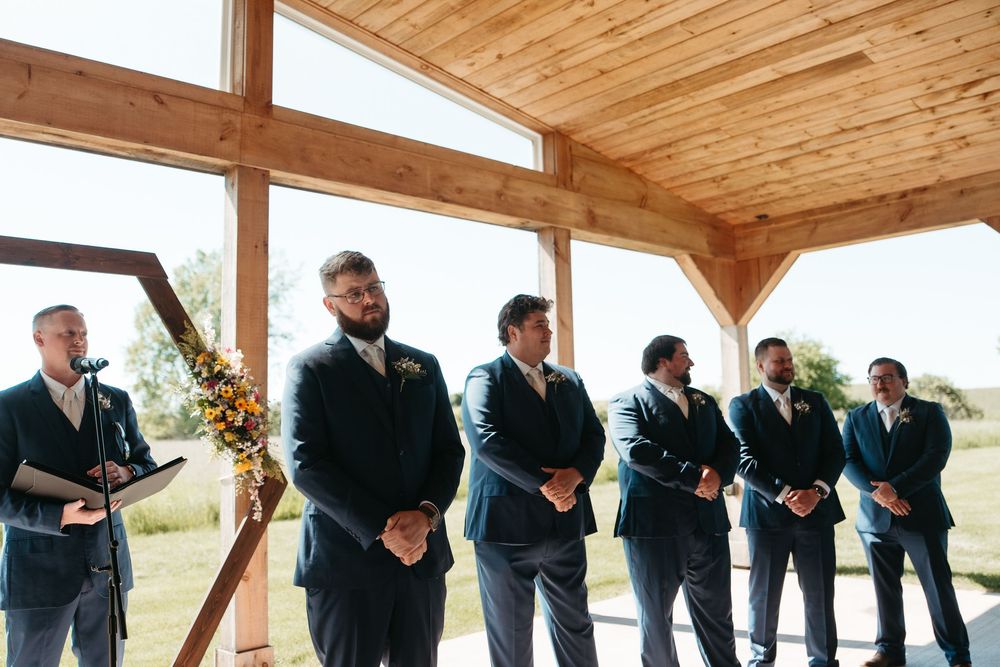 Groom and groomsmen in navy suits stand at wedding altar in rustic wooden venue with large windows.