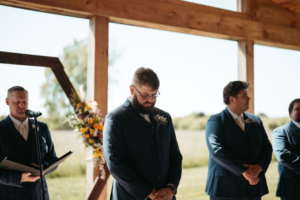 Groom in navy suit bowing head during wedding ceremony in rustic barn venue with groomsmen standing nearby.