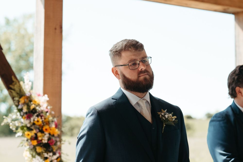 Groom in navy suit with boutonniere and glasses, emotional at wedding altar ceremony.