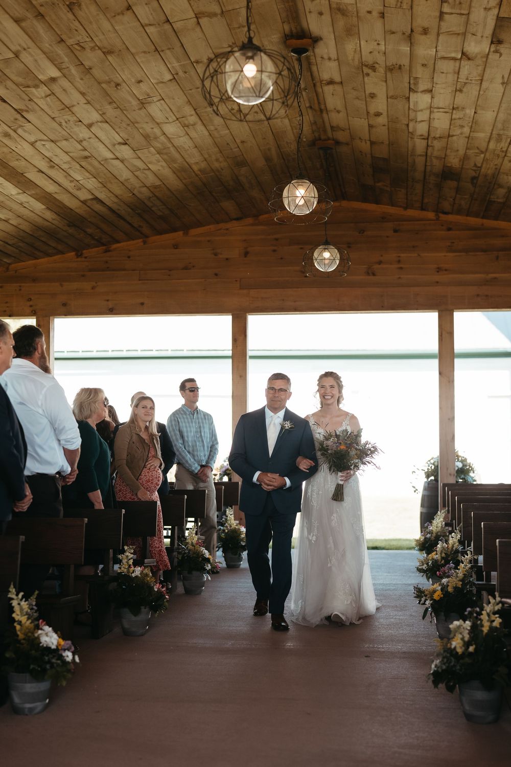 Bride walks down the aisle with escort in a rustic wood chapel, guests standing, floral arrangements lining the aisle.