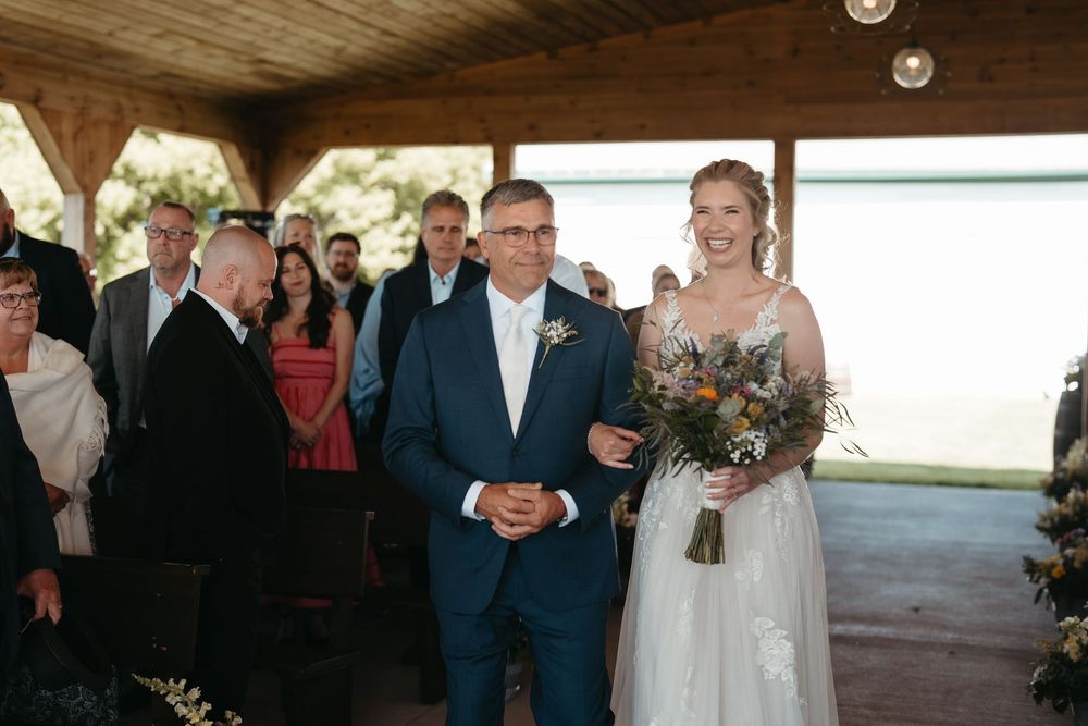 Bride in white gown holding bouquet walks down aisle with escort at outdoor wedding ceremony.