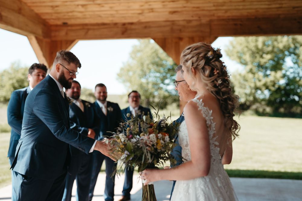 Bride and groom sharing a tender moment during outdoor wedding ceremony, groomsmen standing in background under wooden pavilion.