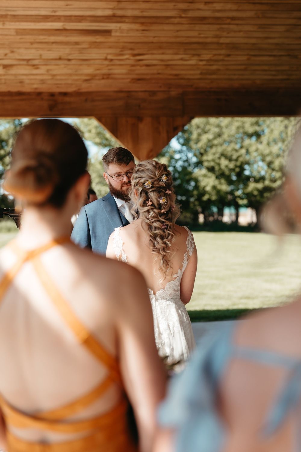 Bride and groom exchange vows under wooden pavilion outdoors, with bridesmaids in yellow and blue dresses watching.