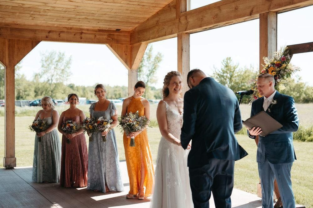 Bride and groom exchange vows under a wooden pavilion with bridesmaids in colorful mismatched gowns holding wildflower bouquets.