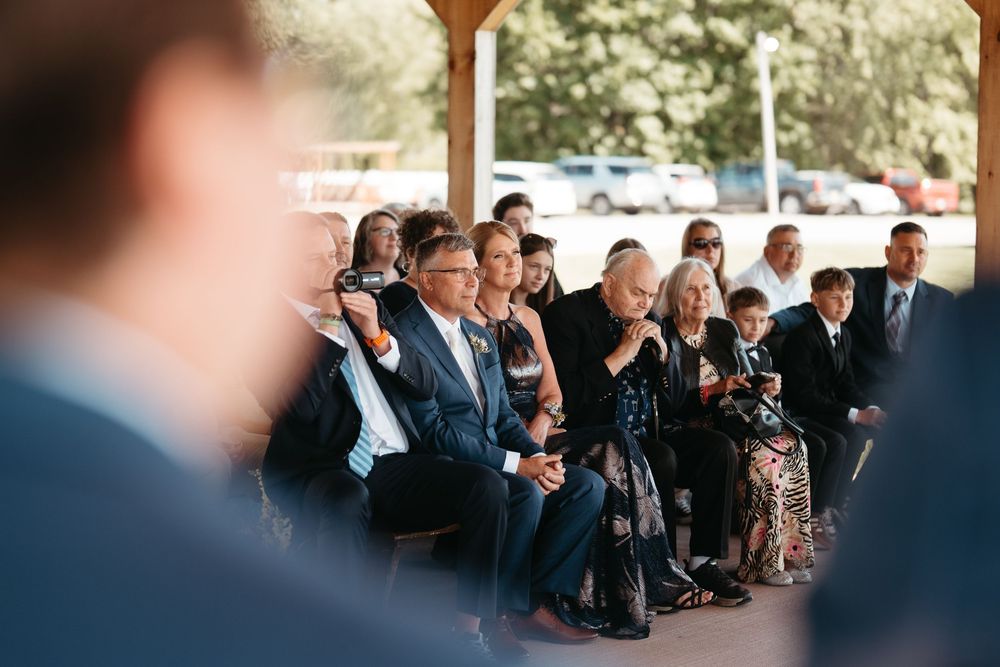 Emotional wedding guests seated under a pavilion, watching an outdoor ceremony on a sunny day.