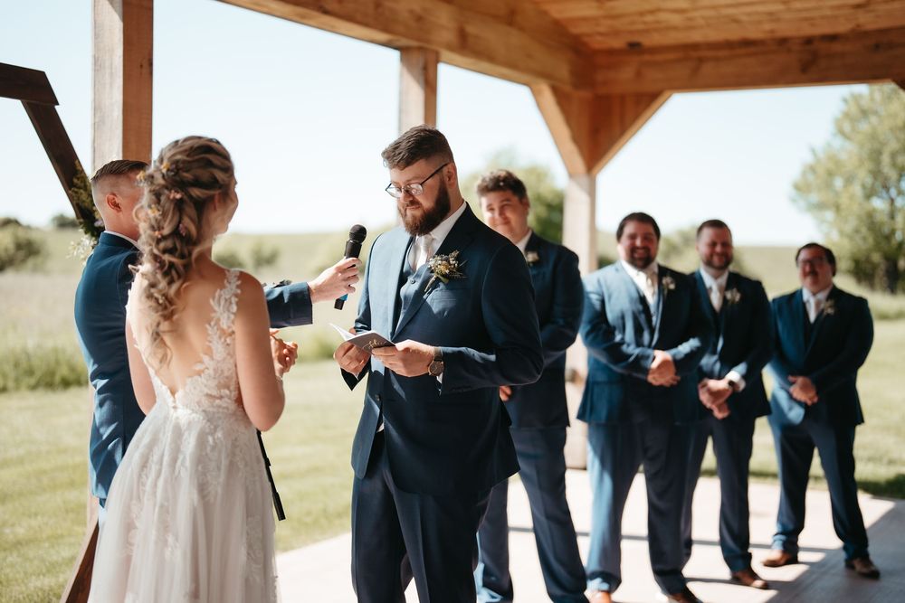 Groom reads wedding vows to bride under rustic pavilion with groomsmen in navy suits standing behind.