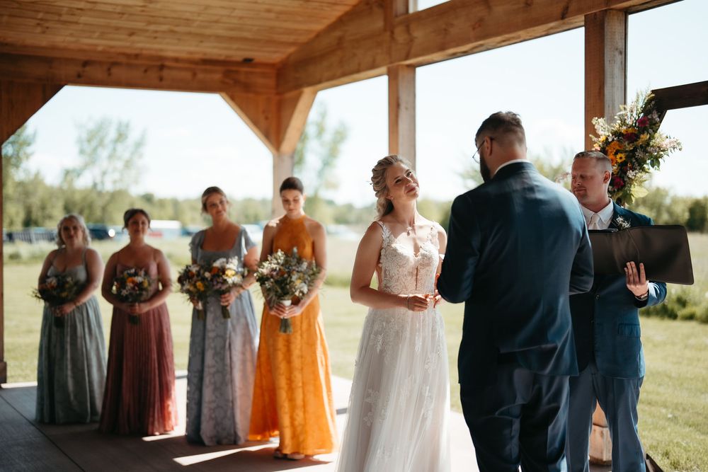 Smiling bride in white gown faces groom at outdoor wedding ceremony with bridesmaids in colorful dresses standing behind.