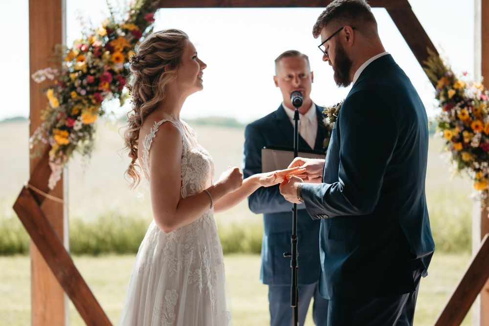Bride and groom exchanging rings at outdoor wedding ceremony under floral arch with officiant.