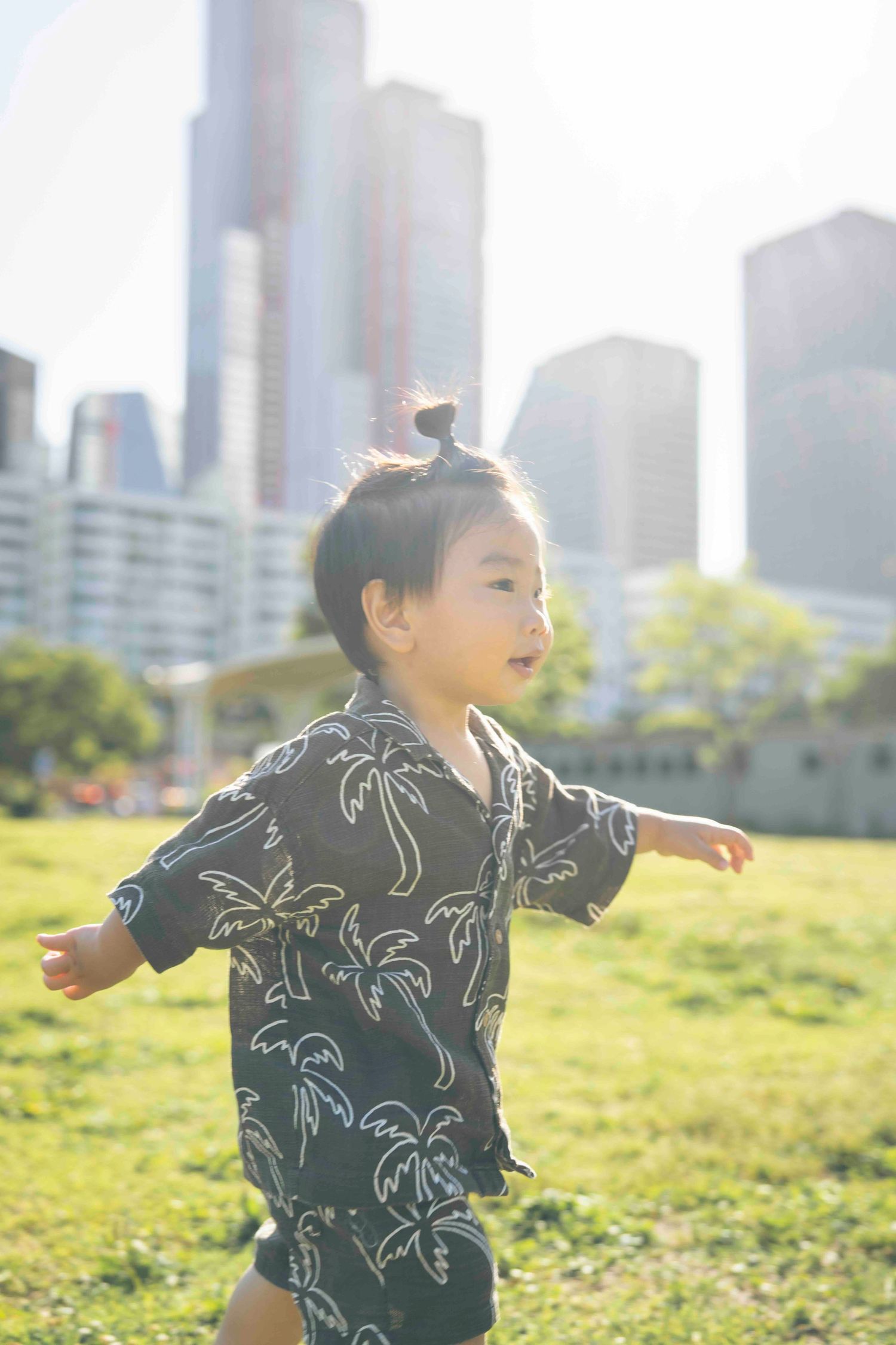 A young Asian toddler in a palm tree print shirt runs joyfully through a green park with city skyscrapers in the background.