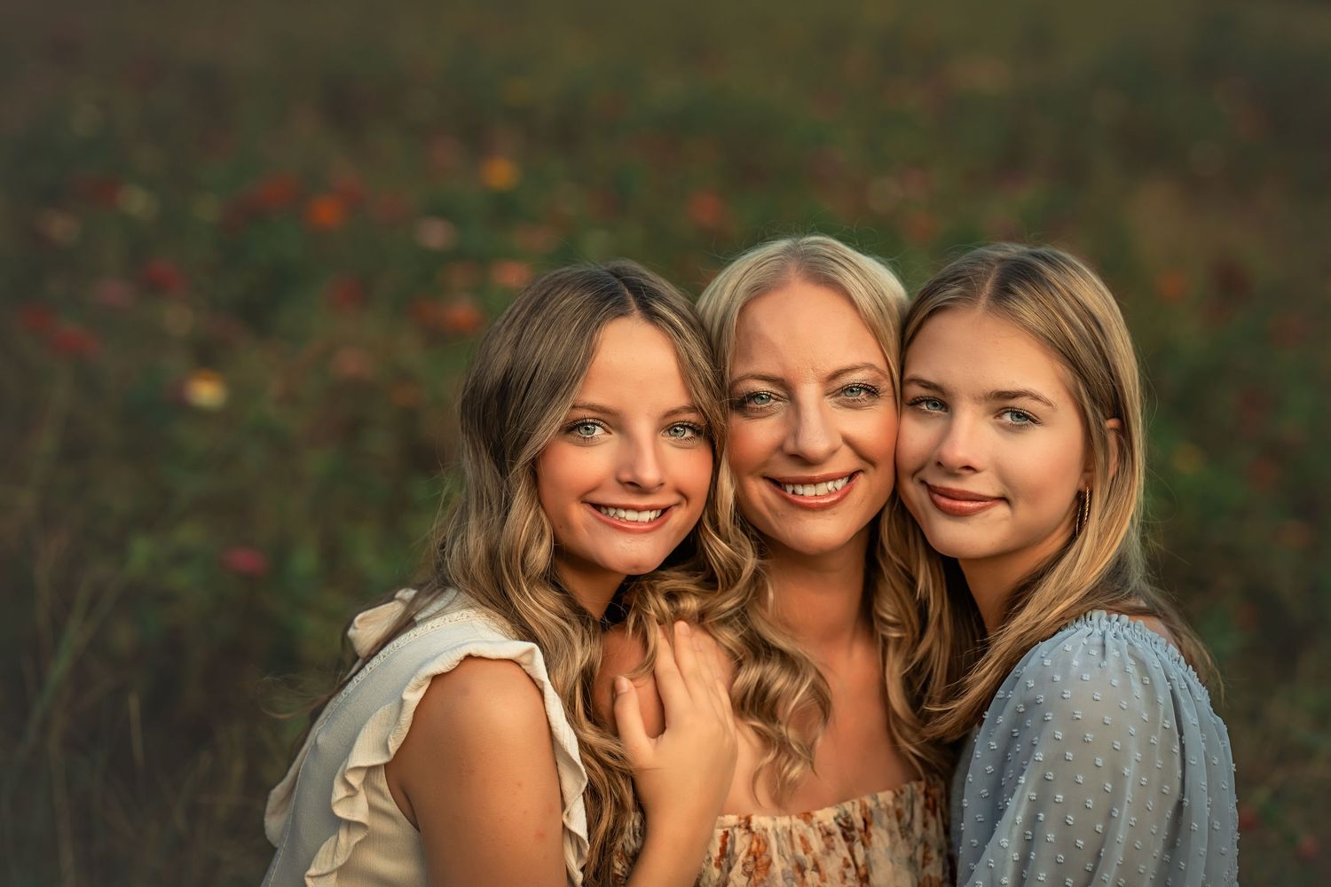 Three smiling blonde women pose together in a sunlit field with colorful wildflowers in the background.