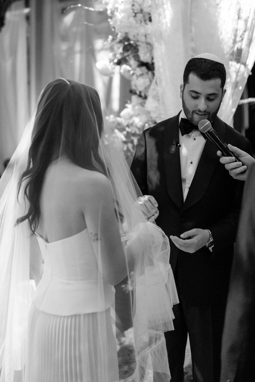 Black and white photo of groom reading vows into microphone to bride in wedding dress and veil at ceremony.