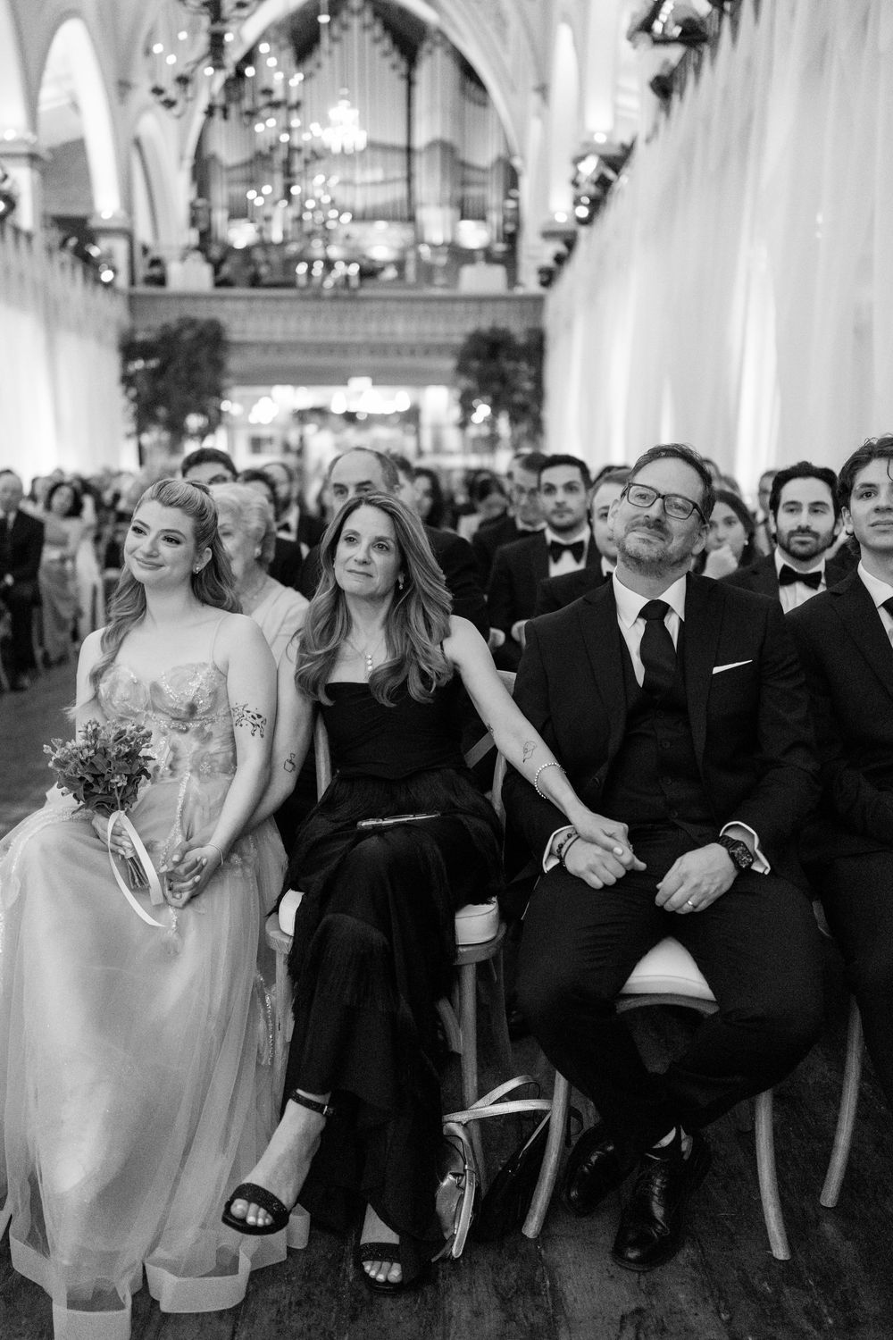 Black and white photo of wedding guests seated in an elegant church, smiling during the ceremony.