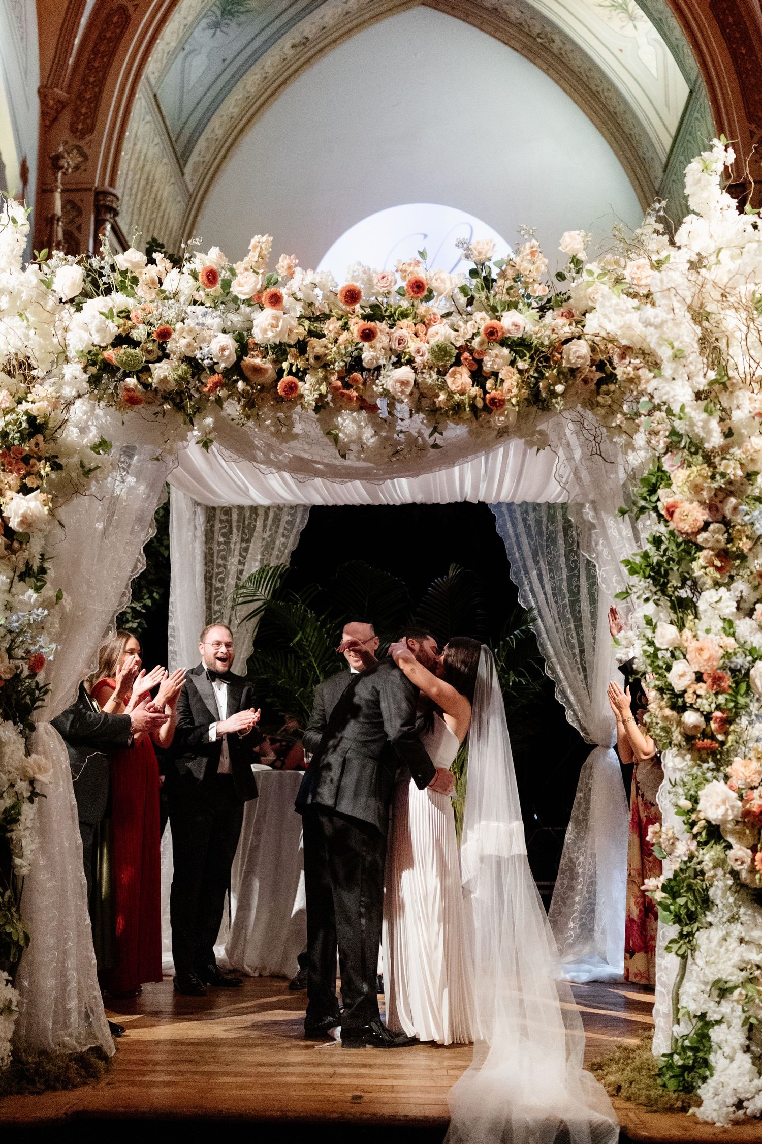 Bride and groom share first kiss under floral chuppah adorned with white and blush roses during elegant wedding ceremony.