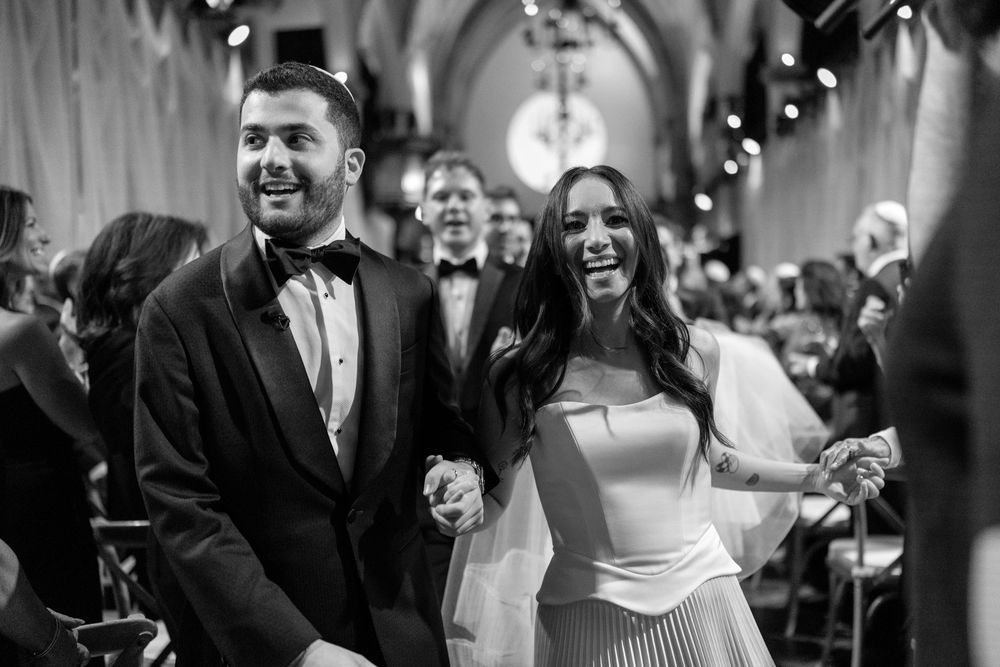 Black and white photo of a joyful couple holding hands walking down the aisle at their elegant church wedding ceremony.