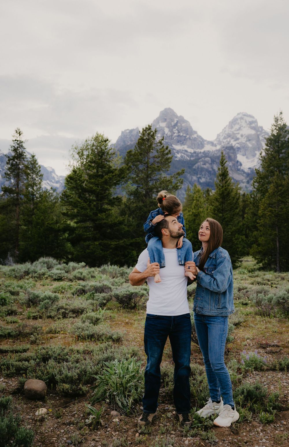 Family of three posing in Grand Teton National Park, with child on father's shoulders and mountains in background.