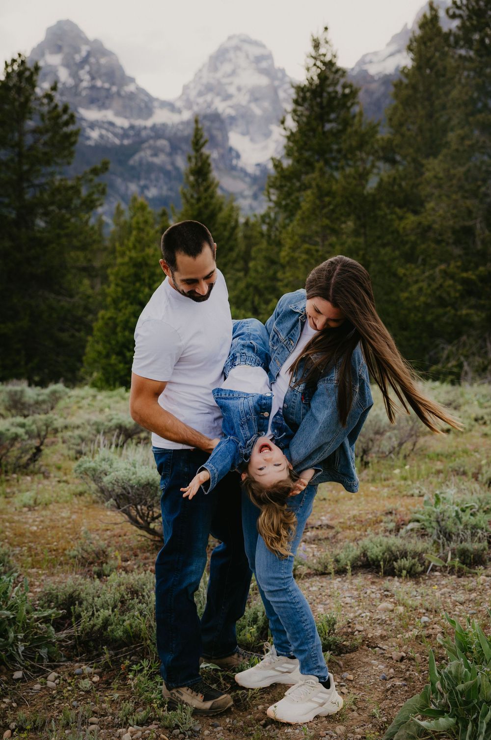 Family of three playfully posing in Grand Teton National Park, parents swinging child upside down among mountain scenery.