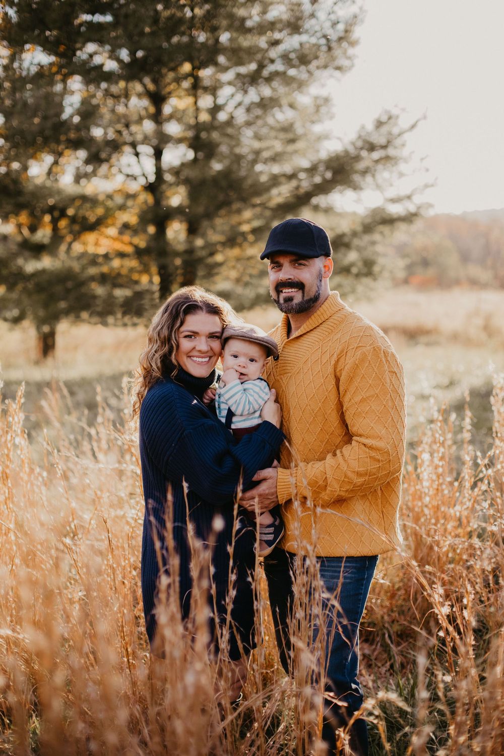Family of three poses in golden autumn field, parents holding baby, father in mustard sweater and cap.