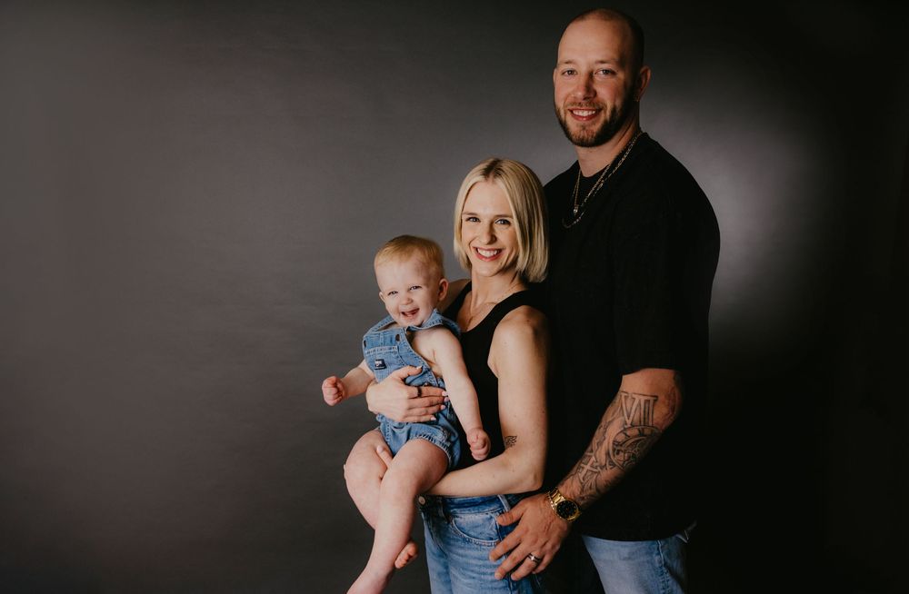 Family portrait of tattooed father, blonde mother, and baby in denim outfit against dark gray studio background.
