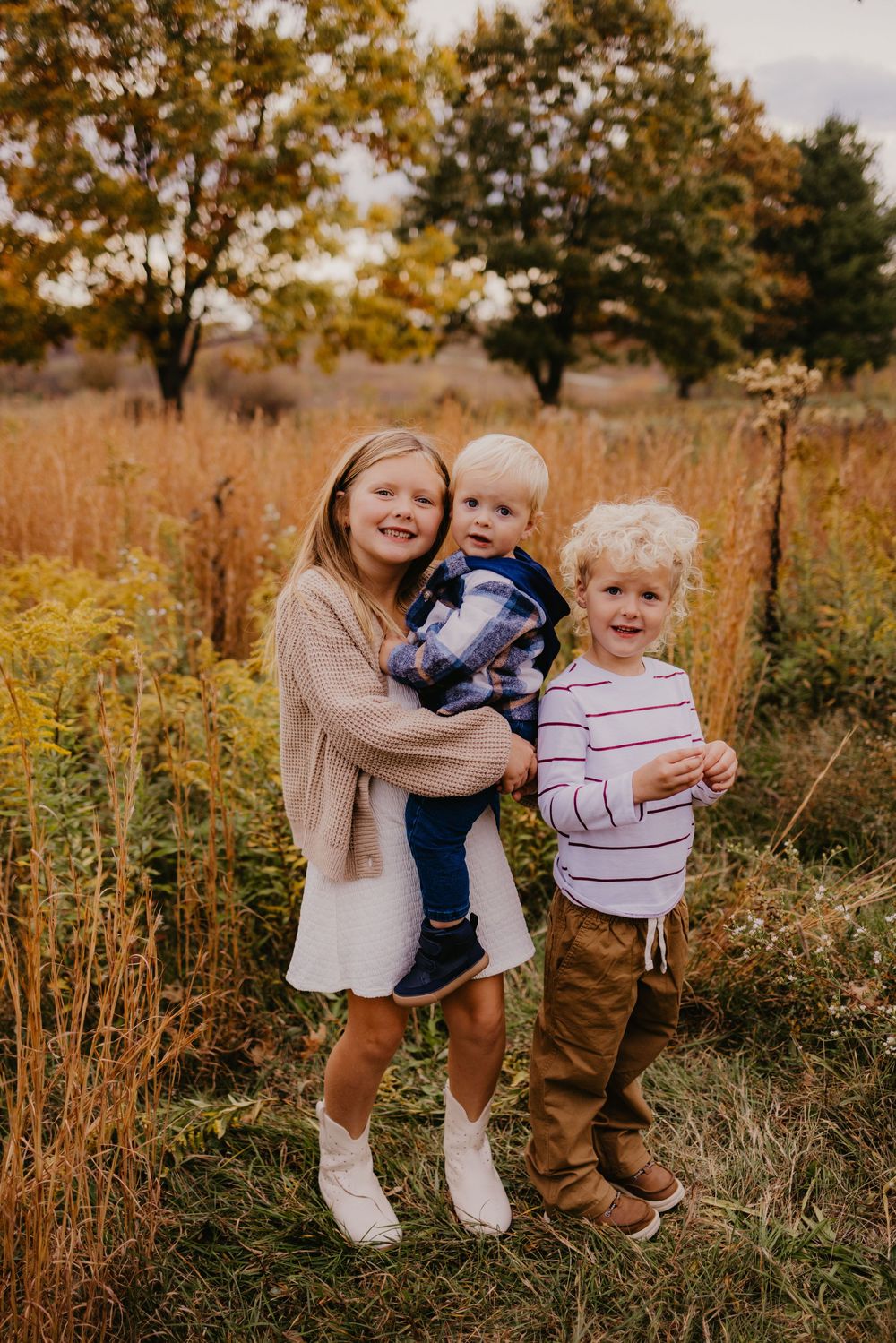 Three siblings pose in autumn field, older girl hugging toddler while young boy stands beside them.