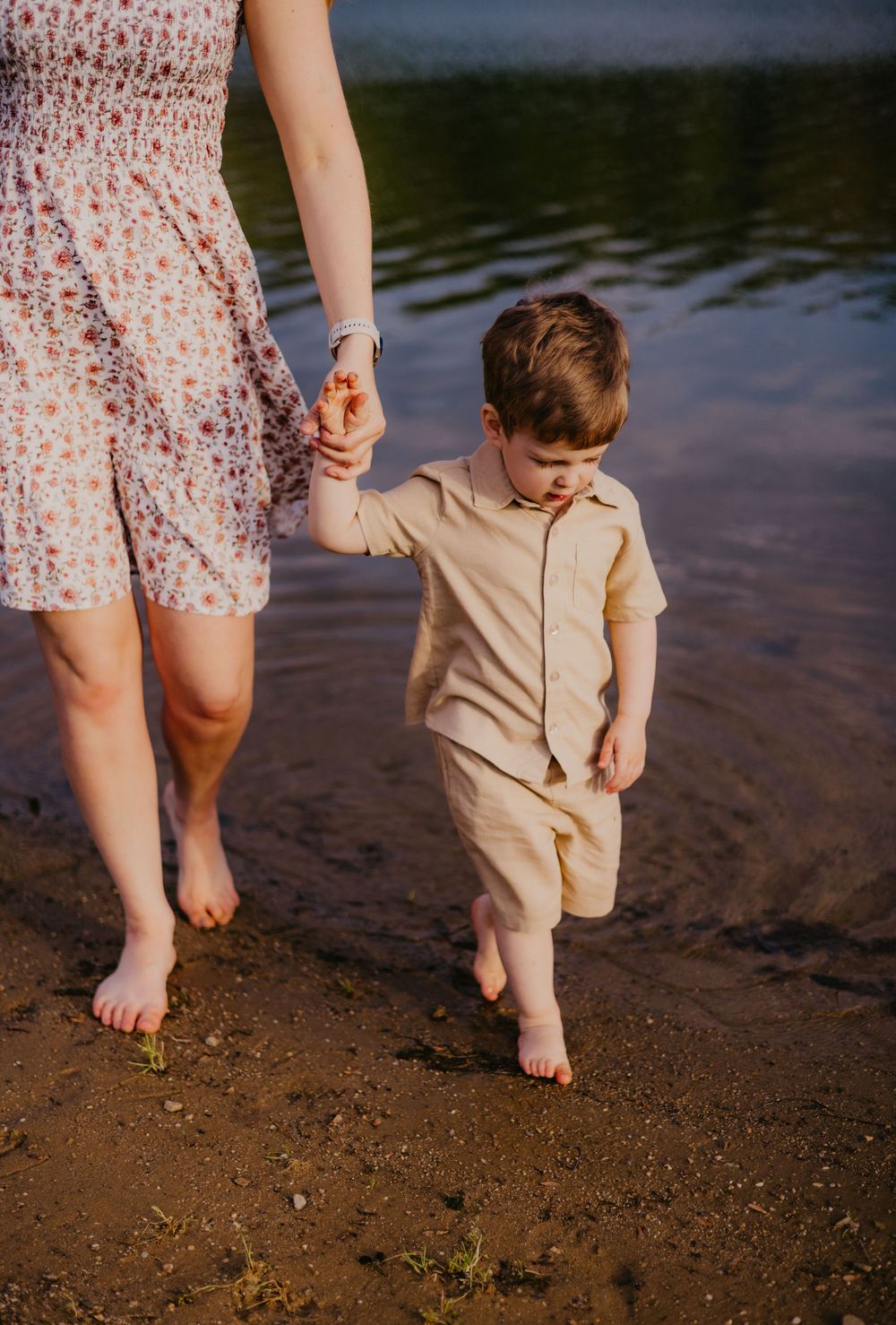 A young boy in a beige outfit holds his mother's hand while wading barefoot along a sandy shoreline.