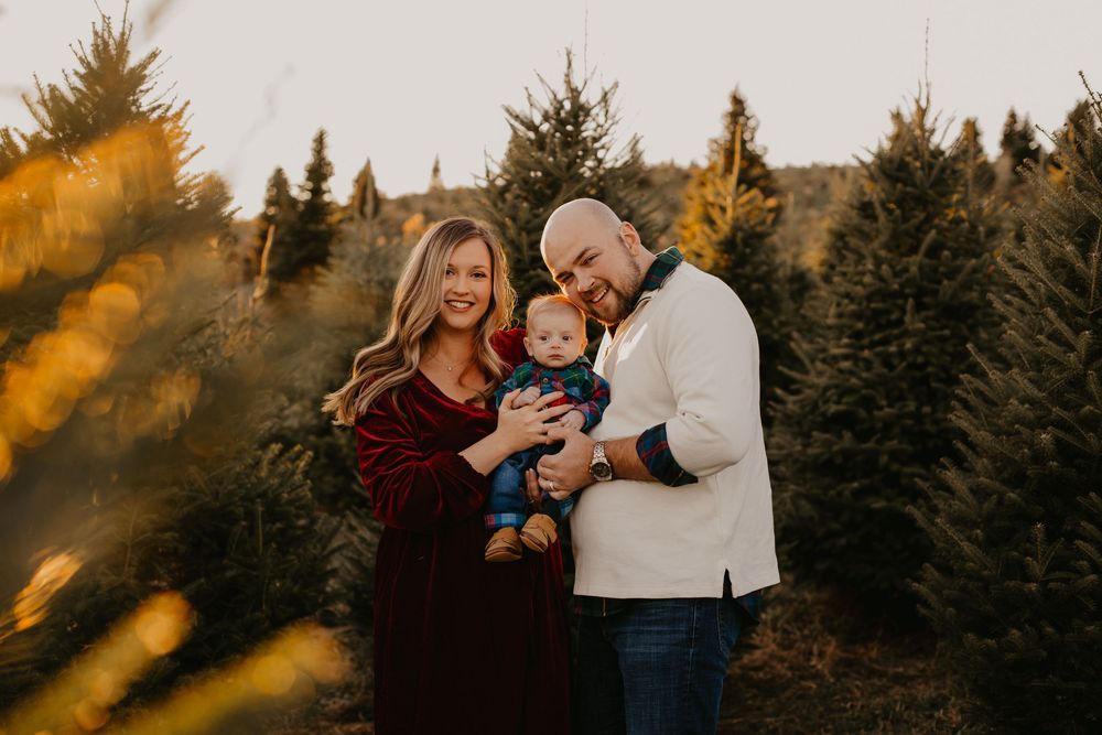 Smiling family with bald father, blonde mother in burgundy dress, and baby at golden sunset tree farm.