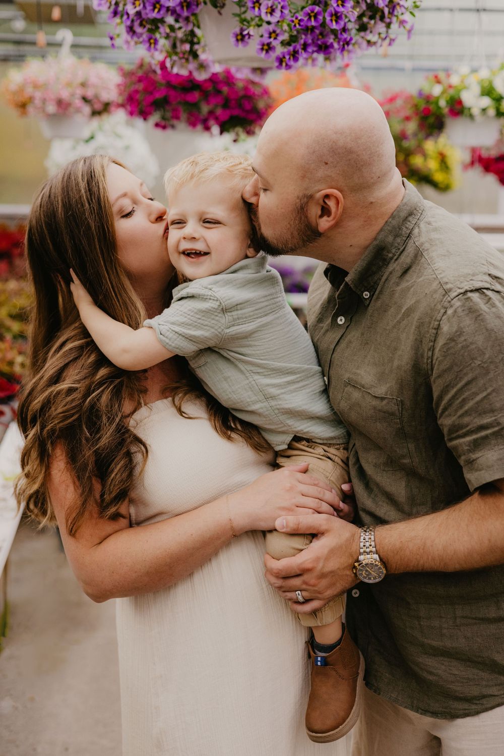 A joyful family portrait with parents kissing their laughing toddler son in a colorful flower greenhouse.