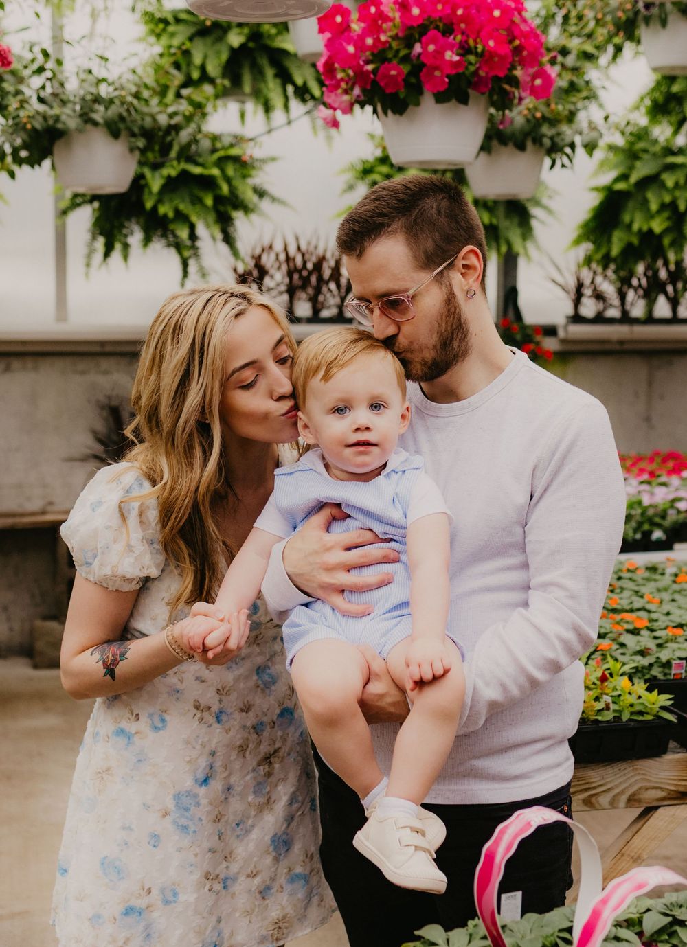 Parents kiss their baby in a floral greenhouse, mother in floral dress and father in white sweater.