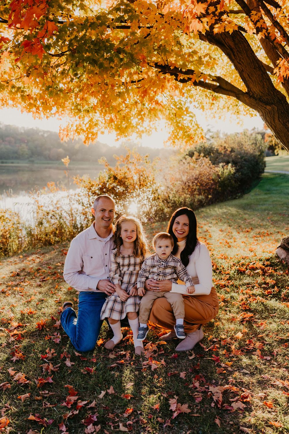 Family of four sits under vibrant orange autumn tree near lake, two young children between parents.
