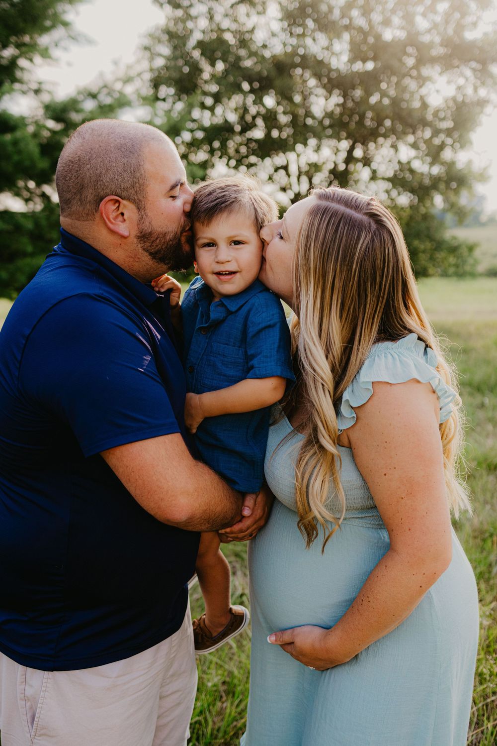 Parents kiss toddler boy on cheeks outdoors at sunset, mother in light blue maternity dress.