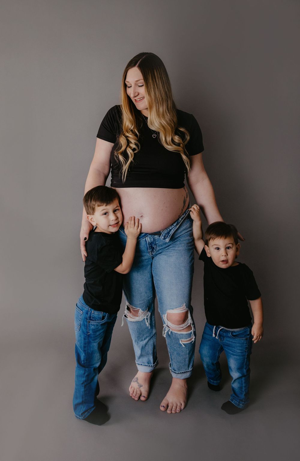 Pregnant woman in black crop top and jeans poses with two young boys hugging her baby bump in a studio maternity photo.