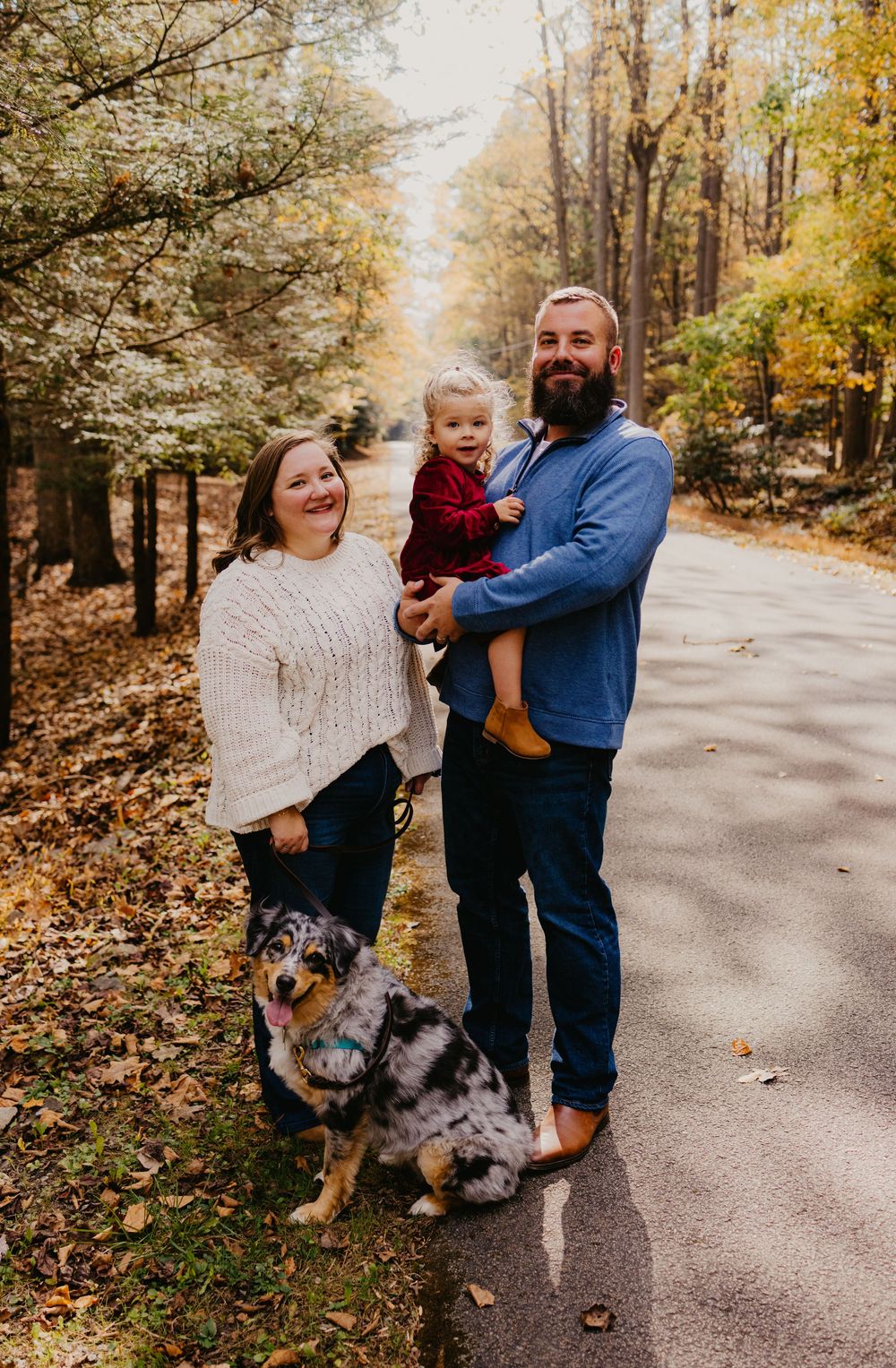 Family with toddler and Australian Shepherd dog posing on a tree-lined road during fall foliage season.