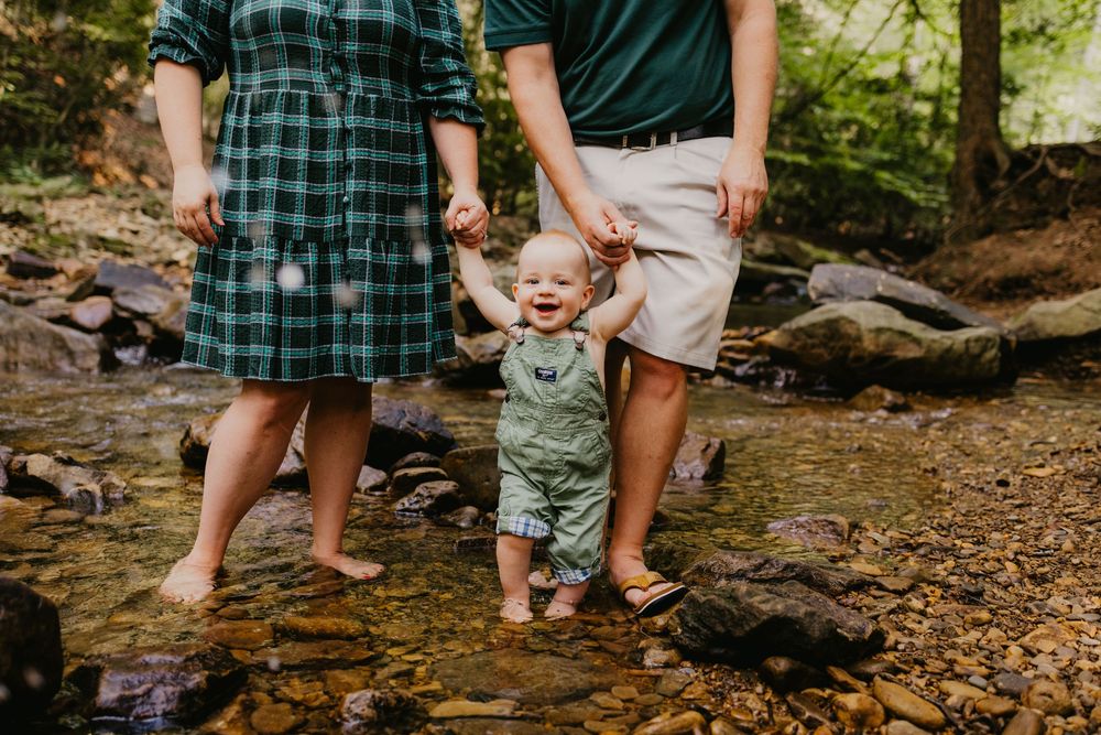 Parents hold baby's hands while wading in a shallow creek during a family photo session outdoors.