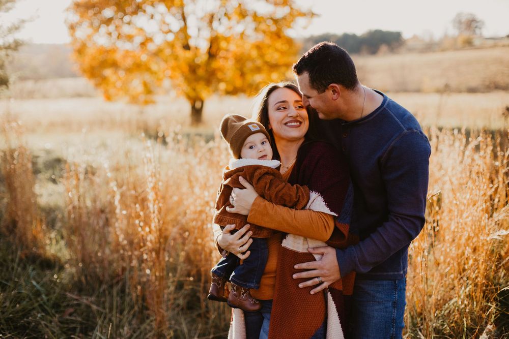Parents embrace their toddler in a golden autumn field at sunset during a family portrait session.
