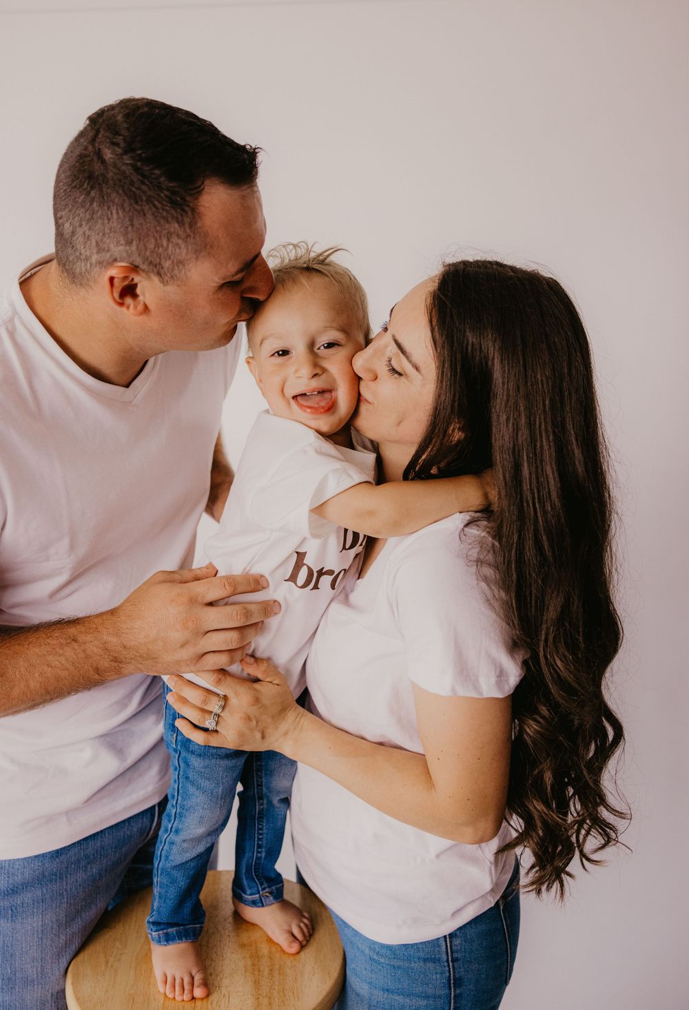 Parents kiss their smiling toddler on the cheeks during a bright studio family photo session.