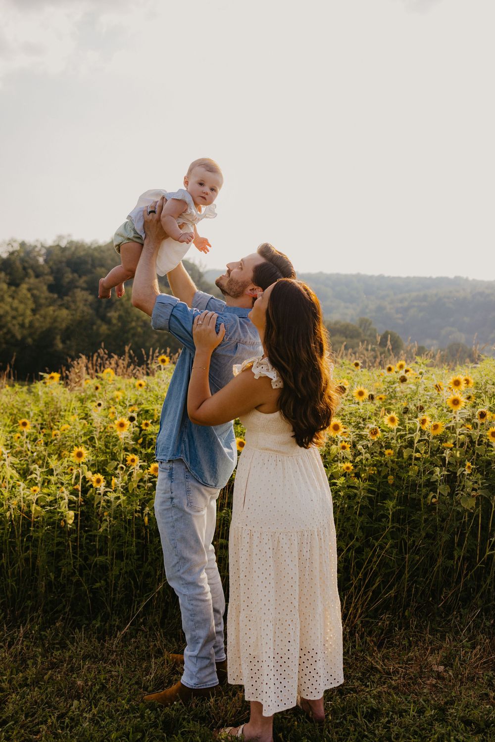 A joyful family in a sunflower field, father lifting baby while mother watches, golden hour light.