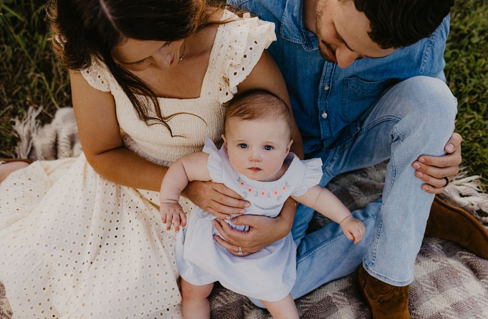 Parents sitting outdoors on a blanket holding their baby girl in a white dress during a family photo session.