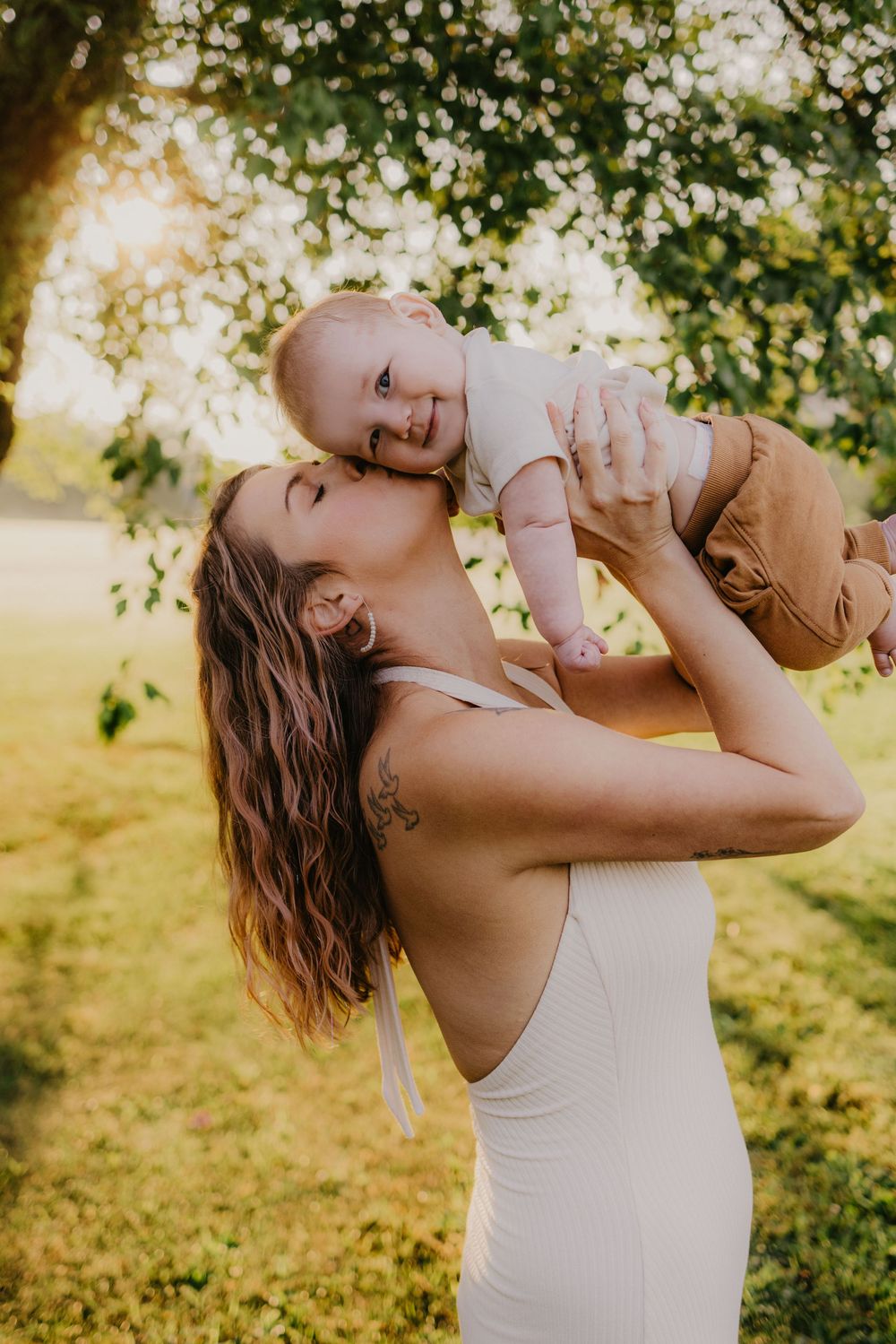 A young mother in a white dress lifts her smiling baby outdoors in golden sunlight beneath green trees.