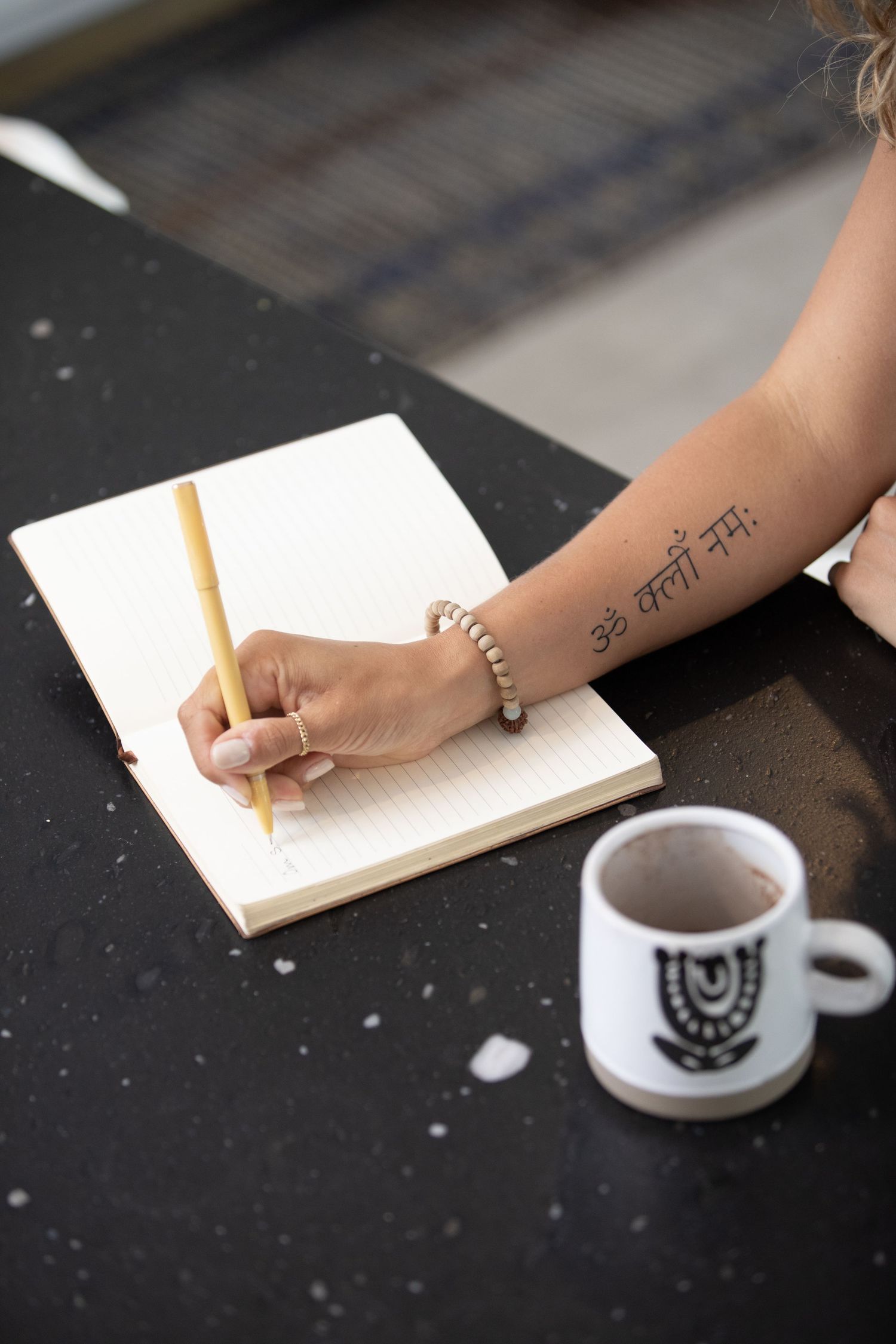 A woman with a Sanskrit tattoo on her forearm writes in a lined notebook with a pencil beside a white mug.