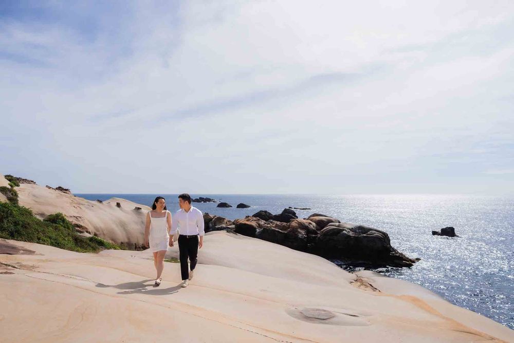 Couple walking hand in hand on sandy coastal rocks with sparkling ocean and blue sky backdrop.