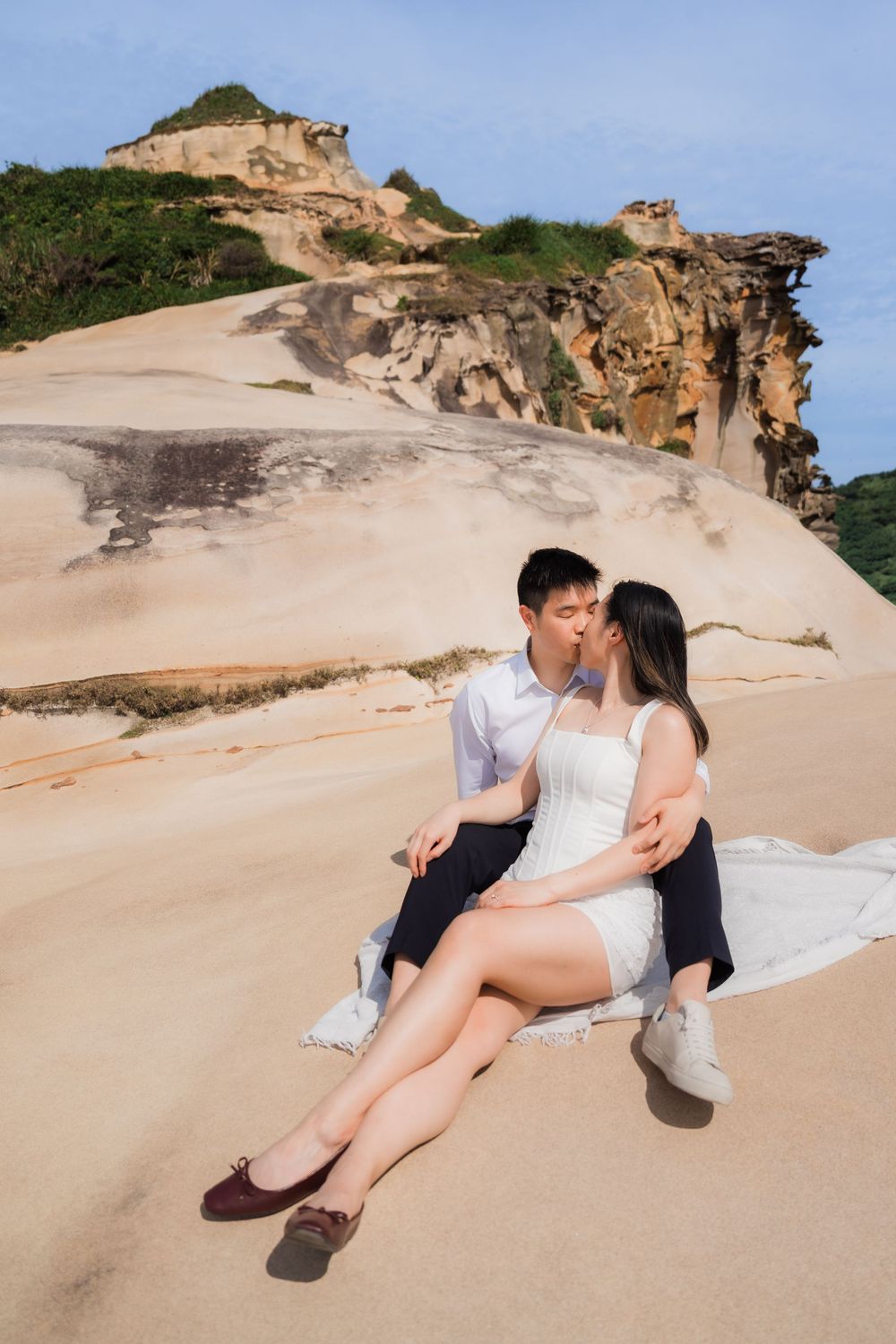 Couple sharing a romantic kiss while sitting on sandy beach rocks with dramatic cliffs and greenery in the background.