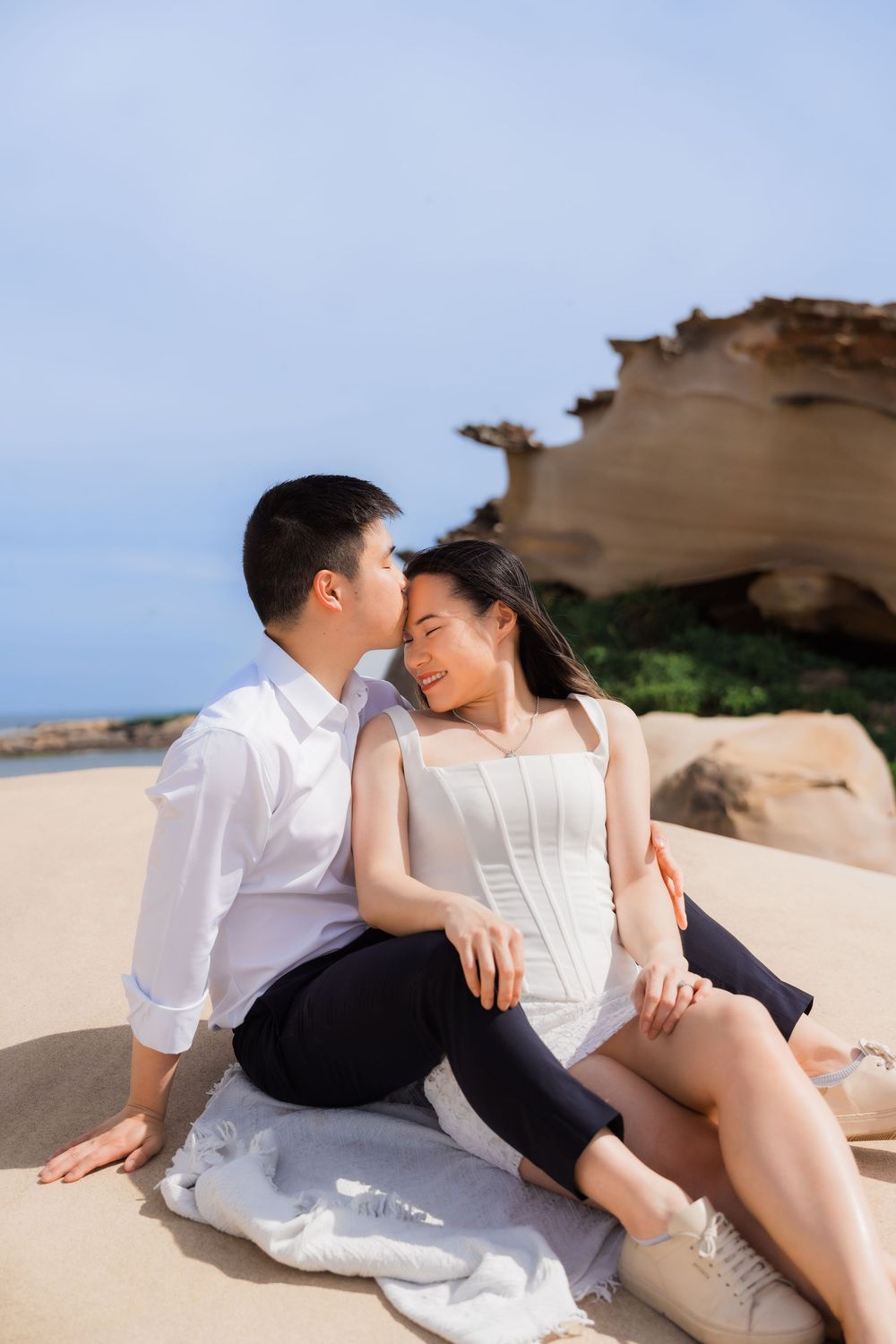 Couple sitting on sandy beach, man kissing woman's cheek, rocky cliffs background, wearing white outfits.