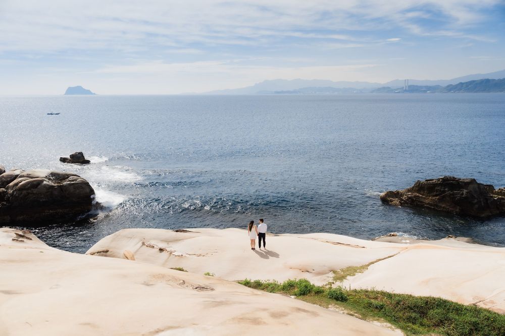 Couple standing on rocky coastal cliffs overlooking calm blue ocean waters with mountains in the distance.